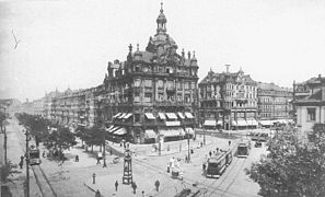 Černobílá fotografie zachycující nároží historizujícího domu s věžičkou oklopeného silnicemi, po kterých jezdí tramvaje